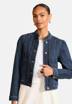 Woman wearing a dark blue denim jacket with scalloped edges, button details, and puffed shoulders over a white top and cream skirt.