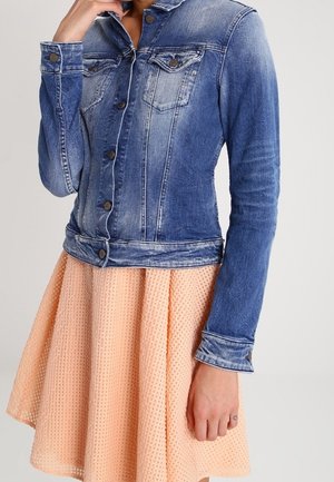 Spijkerjack met knoopsluitingen over een getextureerde perzikkleurige rok, gedragen door een persoon met de ene hand bij het gezicht en de andere langs het lichaam.