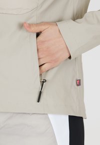 Ljusbeige jacka med en dragkedjeficka, dragsko och elastiska muddar, tillverkad av ett slätt, lätt tyg. Har logotypmärke.