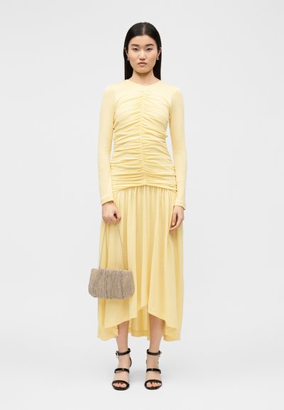 Γυναίκα με κίτρινο μακρυμάνικο φόρεμα με ρυτιδώσεις, κρατώντας μια μπεζ υφασμάτινη τσάντα και φοράει μαύρα σανδάλια με λουράκι στον αστράγαλο.