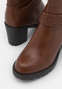 Bruine leren enkellaarsjes met zwarte chunkyzolen engevlochten leren riemen rond de enkels, op een witte ondergrond.