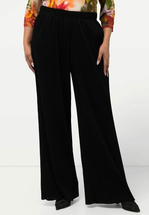 Zwarte, wijde broek met een textuur in ribbelpatroon; elastische tailleband; gedragen met een kleurrijke bloemen blouse met lange mouwen en zwarte schoenen.