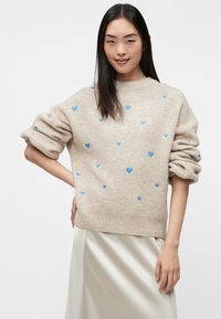 Beige sweater with a loose fit, featuring blue heart patterns and rolled sleeves. The fabric has a soft texture, worn over a light-colored skirt.