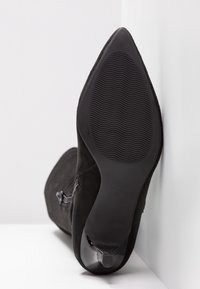 Botte à talon haut en daim noir avec un bout pointu et une semelle en caoutchouc texturée, présentant un accent en cuir lisse sur le talon.