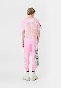 Camiseta de algodón rosa y pantalones de chándal con diseños de alas, con un detalle de etiqueta en los pantalones. Llevado con botas gruesas blancas.