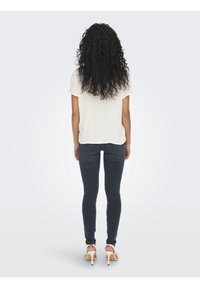 Valkoinen lyhythihainen paita, jossa on teksturoitu kuvio, yhdistettynä tummiin skinny-farkkuihin ja beigen värisiin korkokenkiin. Takakuvassa, hiukset ovat kiharat ja pitkät.