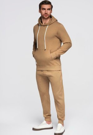 Jonge man die staat met handen in zakken, gekleed in een beige hoodie en bijpassende joggerbroek met witte sneakers, tegen een effen achtergrond.