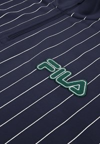 Tummansininen kangas, jossa ohuet valkoiset pystysuorat raidat, varustettuna vihreä-valkoisella FILA-logotarralla ja osittain näkyvällä vetoketjutaskulla.