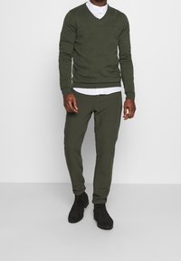 Ciemnozielony sweter typu v-neck wykonany z lekkiego materiału, w połączeniu z dopasowanymi spodniami dresowymi. Slim fit i długie rękawy, z białą koszulką pod spodem.