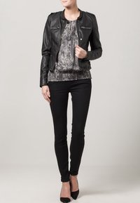 Czarna skórzana kurtka z zamykanymi kieszeniami, zestawiona z szarą wzorzystą bluzką i czarnymi dopasowanymi dżinsami. Modelka ma na sobie czarne szpilki.