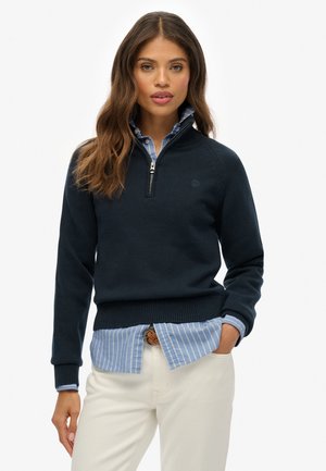 Marineblauwe gebreide trui met een ritssluiting bij de kraag, met geribbelde manchetten en onderzoom, gedragen over een lichtblauw gestreept button-up shirt.