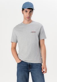 Tricou din bumbac gri, cu mâneci scurte și guler rotund, având un mic logo "GUESS JEANS" roșu și negru pe piept. Purtați cu blugi albaștri și o șapcă albastră.