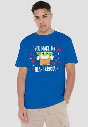 Young man wearing a blue T-shirt with Baby Yoda and "You make my heart Grogu" text surrounded by red hearts and white sparkles.