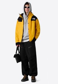 Yellow jacket with black accents, gray hoodie, black pants, and sunglasses. The outfit features a relaxed fit and durable materials.