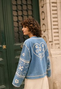 Ženska z kodrastimi lasmi, oblečena v modro jakno z belo vezenim srcem iz cvetličnih vzorcev na hrbtu, stoji ob zelenih vratih in kamnitem zidu.