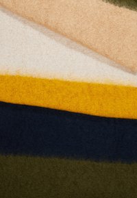 Couches de tissu doux et duveteux en beige, blanc, jaune moutarde, bleu marine foncé et vert olive empilées en diagonale.