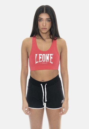 Donna con lunghi capelli scuri che indossa un reggiseno sportivo rosso con il testo "LEONE 1947" e pantaloncini neri con coulisse bianca su sfondo semplice.