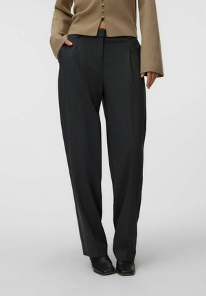 Personne portant un pantalon noir ample à fines rayures, une veste beige courte boutonnée et des chaussures noires à bout carré, debout devant un fond uni.
