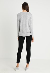 Femme de dos, portant un haut à manches longues gris clair, un pantalon skinny noir, des baskets argentées et des boucles d'oreilles créoles sur fond blanc.