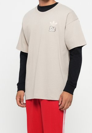 Personne portant une chemise beige à manches courtes sur des manches longues noires avec un petit logo blanc et un pantalon rouge avec des rayures blanches.