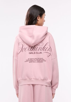 Donna con capelli lunghi e scuri che indossa una felpa con cappuccio rosa e pantaloni abbinati, mostra il testo sul retro "Overthinking Girls Club" e una citazione stampata.