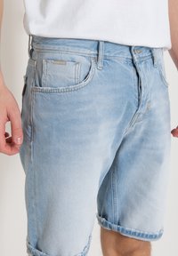 Pantalones cortos de mezclilla azul claro con un acabado desgastado, dobladillo enrollado, bolsillos frontales y una pequeña etiqueta con el logotipo cerca de la cintura.