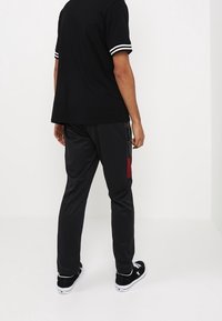 Mies, joka on pukeutunut mustaan polo-paitaan ja mustiin verkkohousuihin, joissa on punaisia yksityiskohtia, seisoo valkoista taustaa vasten ja kääntää selkänsä kameraan.