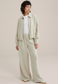 Svetle béžová bomber bunda s elastickými manžetami, kombinovaná s volnými béžovými kalhotami. Model má na sobě bílou košili a světlé boty. Hladká textura.