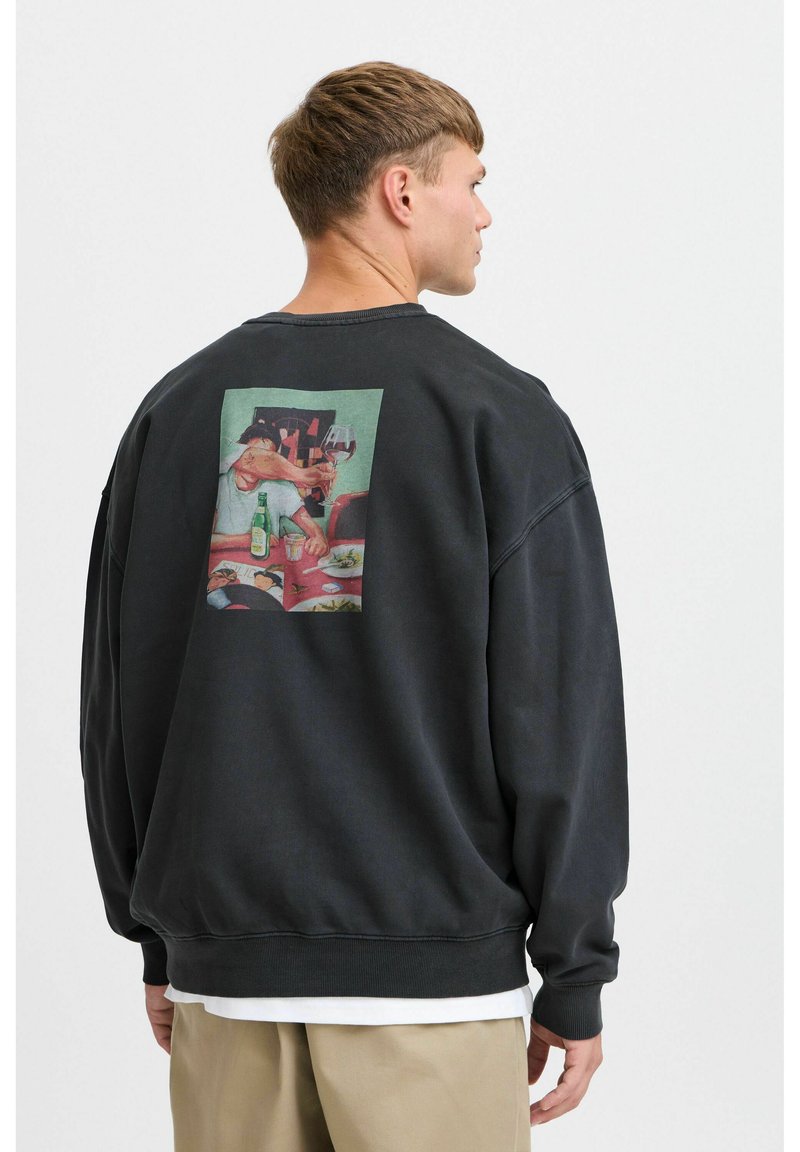 Svart genser med løs passform, med et rektangulært motiv på baksiden som viser en spisescene med en flaske og et glass.