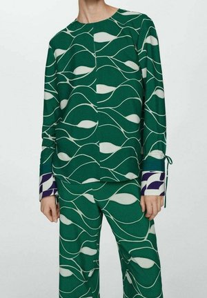 Persona vistiendo conjunto verde de camiseta de manga larga y pantalones con patrones abstractos de olas blancas y detalles en los puños en contraste morado y blanco.