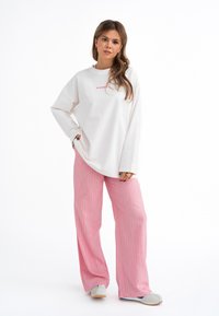 Felpa bianca a maniche lunghe con testo rosso, abbinata a pantaloni a gamba larga a righe rosa chiaro e scarpe grigie con accenti blu.