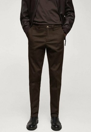 Mand iført mørkebrune skræddersyede bukser, sort læderjakke og sorte læderstøvler, stående med hænderne i lommerne.