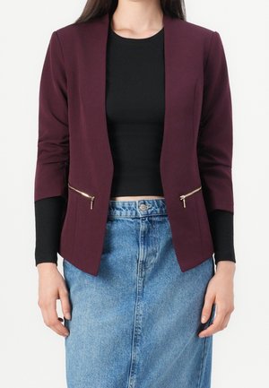 Femme portant un crop top noir, un blazer bordeaux avec des poches zippées dorées et une jupe en jean bleue taille haute sur un fond uni.