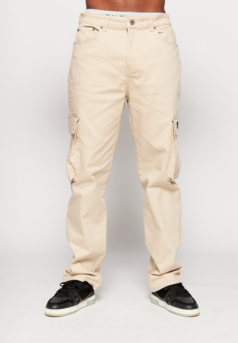 Pantaloni cargo beige in cotone, con tasche laterali, gambe dritte e chiusura con bottone. Indossati con scarpe sportive nere.