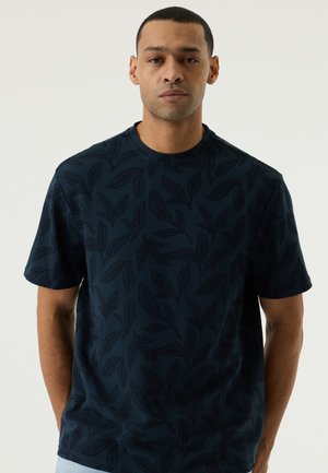 Man die naar voren kijkt en een donker marineblauw T-shirt met korte mouwen draagt met een subtiel bladmotief op een witte achtergrond.