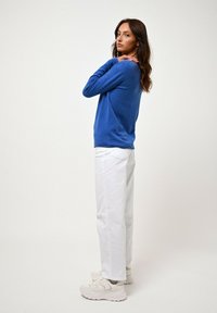 Blå stickad tröja, vita byxor och kraftiga vita sneakers. Tröjan har lång ärm och en avslappnad passform. Byxorna är rakbenade.