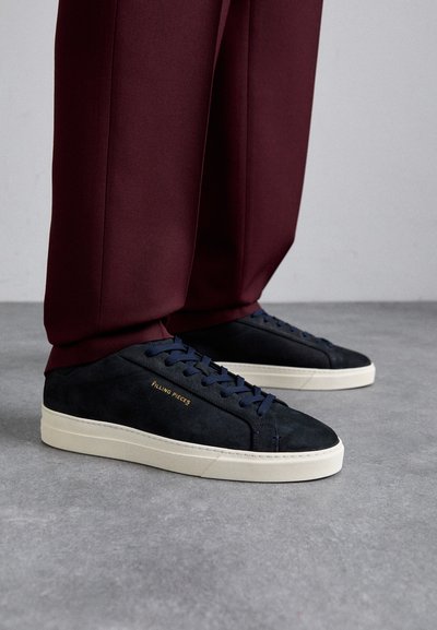 Jūras spēku zila zamša sneakers ar bēšiem zolēm, kura ir aprīkota ar jūras spēku ziliem šņorēm un "Filling Pieces" zeltītu logo. Saskaņoti ar bordo izdotiem biksēm.