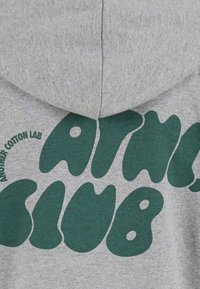 Szürke kapucnis pulóver, a hátán merész, szabálytalan zöld betűkkel "ANOTHER COTTON LAB CLUB" felirattal, puha, textúrájú anyagból.