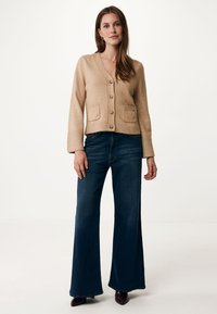 Beige stickad cardigan med V-ringning, knäppning och två framfickor. I kombination med mörkblå utsvängda jeans och mörkbruna skor.