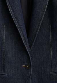 Blazer en denim bleu foncé avec surpiqûres beige, un bouton noir unique et deux poches frontales horizontales.