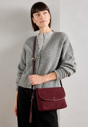 Bordeauxfarbene Wildleder-Schultertasche mit Klappenverschluss, ausgestattet mit einem Quasten-Detail und goldenen Akzenten. Gewebte graue Strickjacke über einem hellen Oberteil getragen.