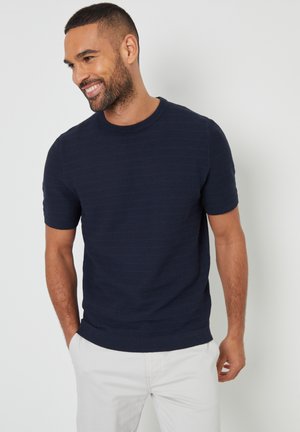 Pull en tricot à manches courtes bleu marine avec un motif texturé et un col rond côtelé, associé à un pantalon de couleur claire.