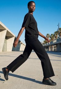 Kvinde iført sort T-shirt, sorte bukser med vide ben og sorte sneakers går udendørs på en solskinsdag nær moderne betonbygninger og palmer.