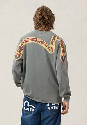 Uomo con capelli mossi di media lunghezza indossa una maglietta grigia a maniche lunghe con motivo fiamma giallo-rosso e jeans blu con loghi bianchi "EVISU" sulle tasche.