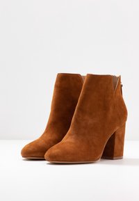 Pari nahkasaappaa, ruskeita mokkasaappaita, joissa on paksut korot ja huomaamaton takavetoketju valkoista taustaa vasten.