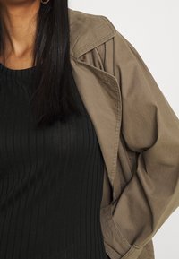 Beige, übergroße Jacke mit einem strukturierten Kragen, kombiniert mit einem schwarzen Rippoberteil. Die Materialien wirken strukturiert mit sichtbaren Nähten.