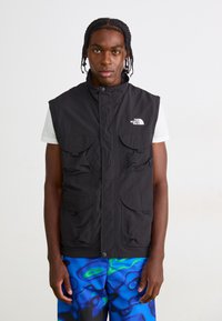 Ung mand med fletninger iført en sort ærmeløs North Face vest over en hvid t-shirt og blå mønstrede bukser, stående mod hvid baggrund.