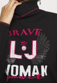 Pull à col roulé noir avec le texte audacieux "BRAVE LJ WOMAN", orné d'accents en strass et de surlignages roses le long du col.