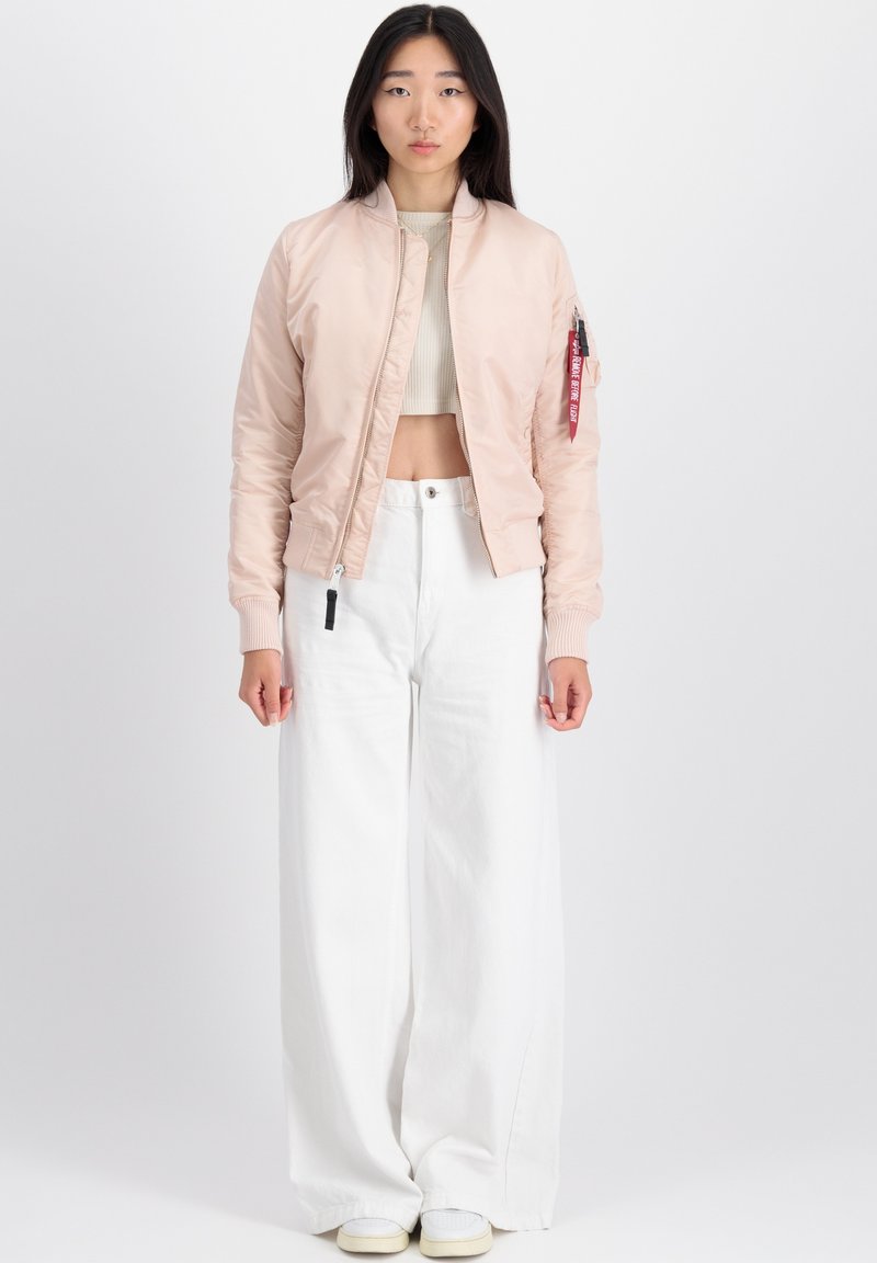 Helles rosa Bomberjacke mit einem cropped Design, gerippten Bündchen und einem Reißverschluss. Mit einem roten Etikett am linken Arm. Kombiniert mit weißen weiten Hosen.