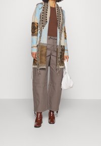 Mönstrad, flerfärgad stickad cardigan med fransar, blå och brun design; brun ribbad topp; grå vida byxor; glänsande vinröda anklarstövlar; vit väska.